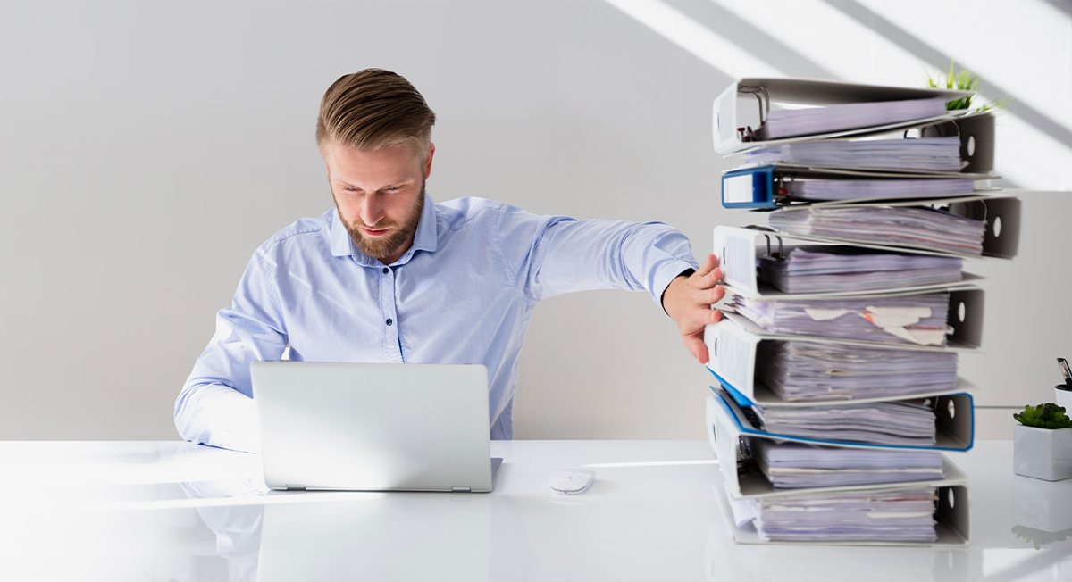 Mann sitzt am Laptop und hat einen Aktenstapel neben sich auf dem Schreibtisch liegen.