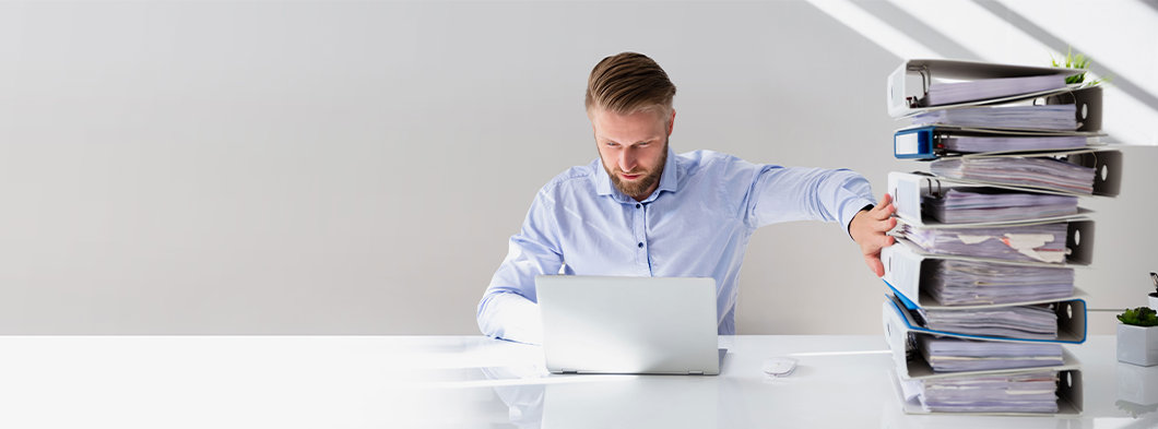 Mann sitzt am Laptop und hat einen Aktenstapel neben sich auf dem Schreibtisch liegen.