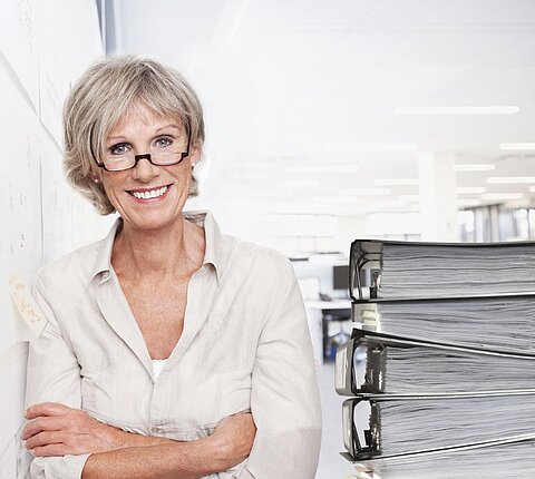 Frau mit Brille, lächelnd in einer Büroumgebung, stehend neben einem Stapel Aktenordner